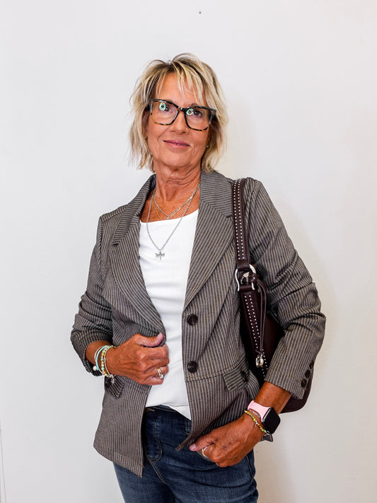 Woman wearing a brown  blazer, white shirt, and jeans with a handbag against a plain background