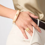 Hand wearing a gold ring and beige bracelet on a neutral background