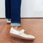 A person wearing off-white slip-on sneakers with white side stripes and a hook-and-loop strap, paired with cuffed dark blue jeans, standing on a terrazzo-patterned floor.