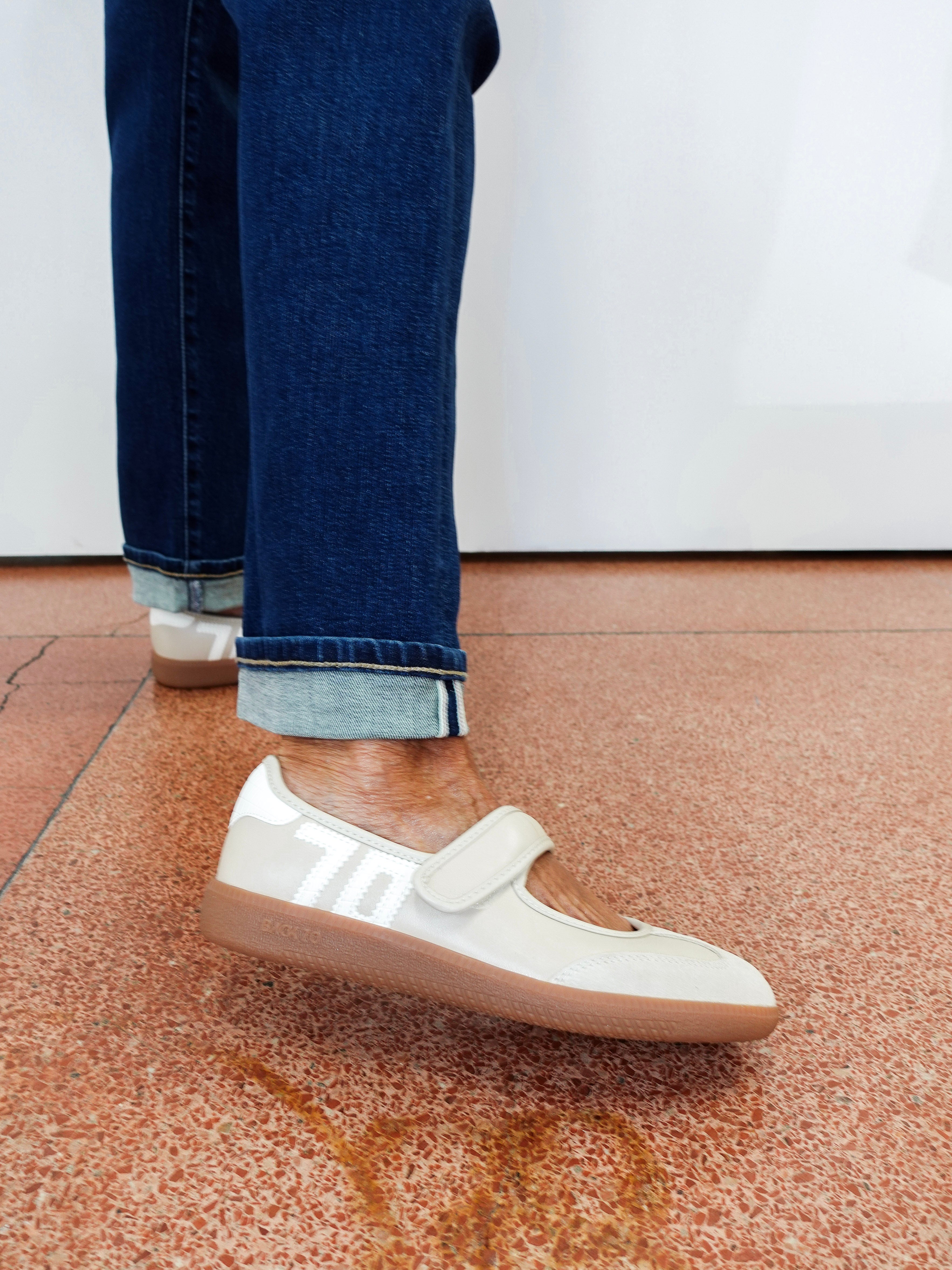 A person wearing off-white slip-on sneakers with white side stripes and a hook-and-loop strap, paired with cuffed dark blue jeans, standing on a terrazzo-patterned floor.