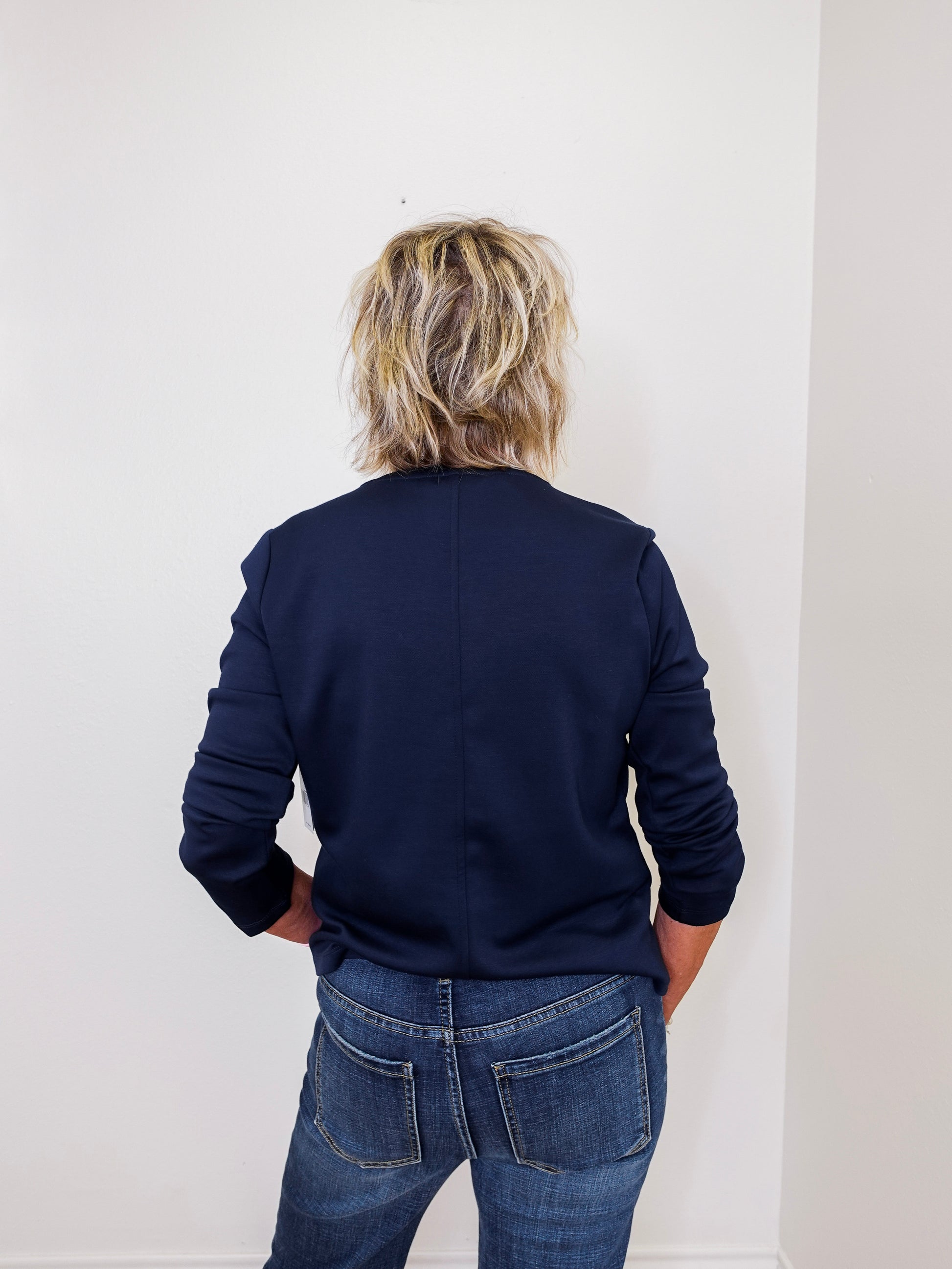 Person wearing a navy blue top and jeans standing against a white wall.