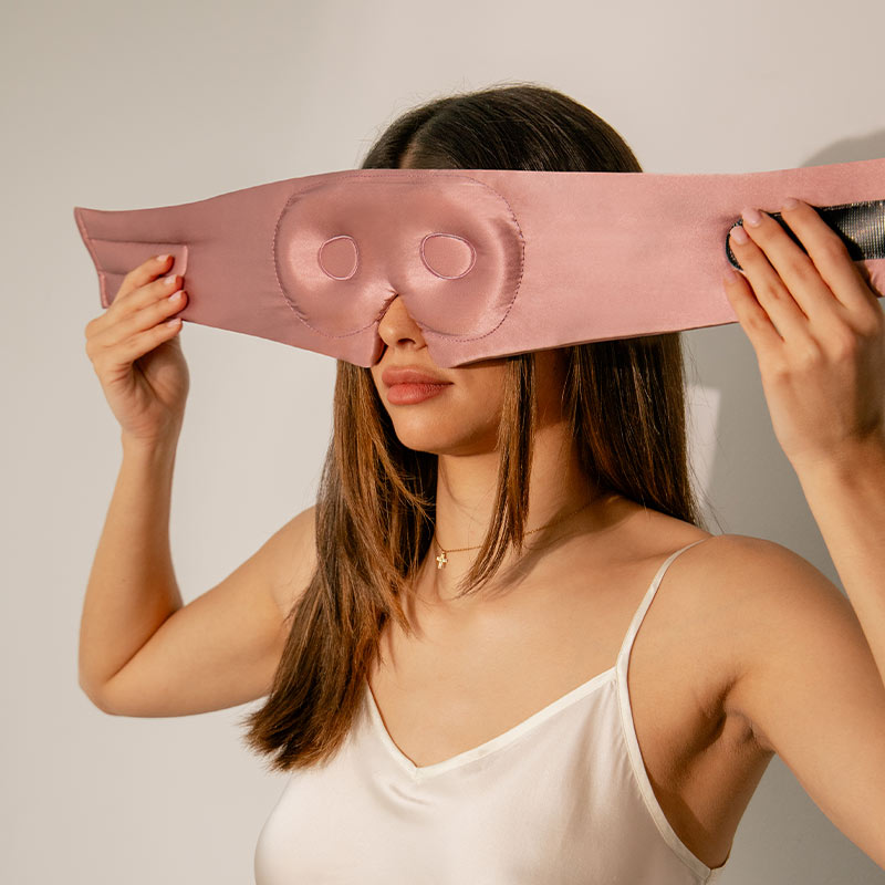 Woman holding a pink satin eye mask over her eyes against a plain background