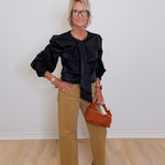 Woman wearing a black blouse with ruffled sleeves and tan pants, holding a brown handbag.