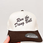 White and brown cap with 'Run the Dang Ball' text held by a hand on a light gray background