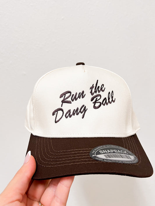 White and brown cap with 'Run the Dang Ball' text held by a hand on a light gray background