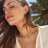 Woman wearing gold heart-shaped earrings and necklace with a blurred outdoor background