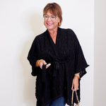 Woman wearing a black poncho and holding a patterned handbag against a white wall.