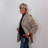 Person wearing a khaki blazer over a black top and blue jeans against a white wall.