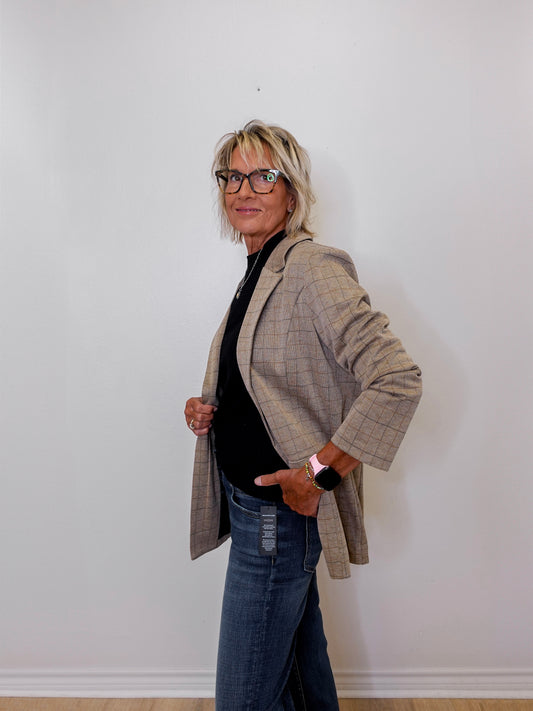 Person wearing a khaki blazer over a black top and blue jeans against a white wall.