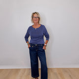 Woman wearing a navy top and blue jeans standing against a plain white wall.