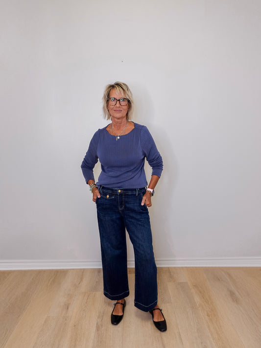 Woman wearing a navy top and blue jeans standing against a plain white wall.