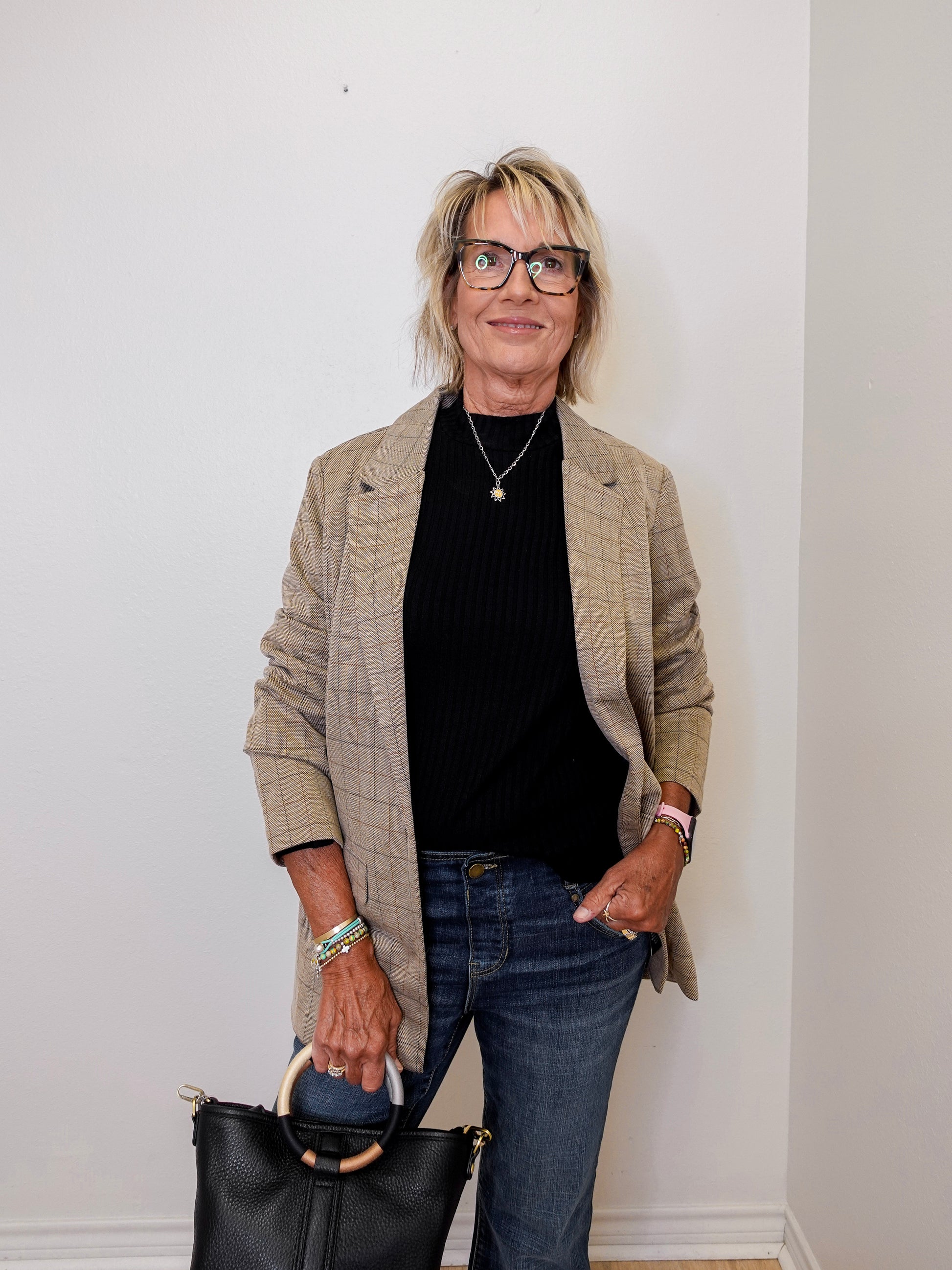 Woman wearing a plaid blazer, black top, and jeans, holding a black handbag against a white wall.