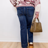 Person wearing a patterned top, blue jeans, and brown shoes, holding a green handbag against a white wall.