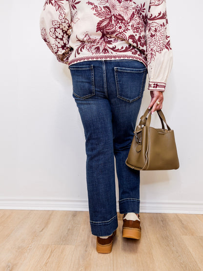 Person wearing a patterned top, blue jeans, and brown shoes, holding a green handbag against a white wall.