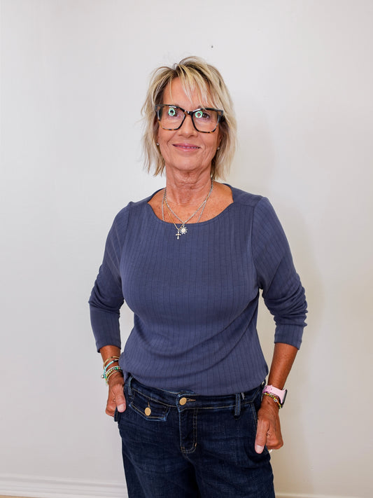 Woman wearing a blue long-sleeve top and dark jeans against a white wall