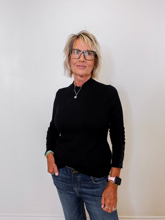 Person wearing a black ribbed sweater and blue jeans against a white wall