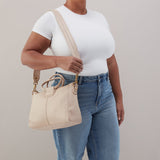 Person holding a beige handbag wearing a white shirt and blue jeans against a plain background