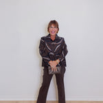 Woman wearing a patterned jacket and dark pants against a white wall.
