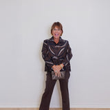 Woman wearing a patterned jacket and dark pants against a white wall.