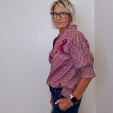 Woman wearing a patterned blouse and jeans against a plain wall.