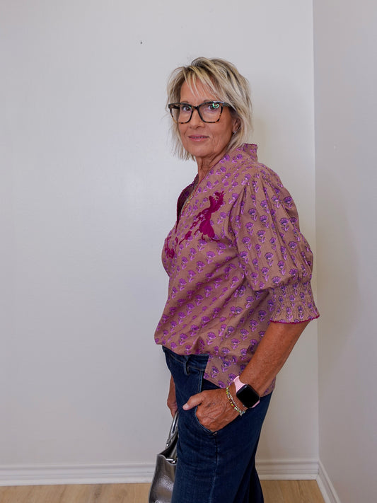 Woman wearing a patterned blouse and jeans against a plain wall.