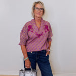 Woman wearing a patterned shirt and jeans, holding a silver handbag against a white wall.