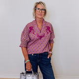 Woman wearing a patterned shirt and jeans, holding a silver handbag against a white wall.