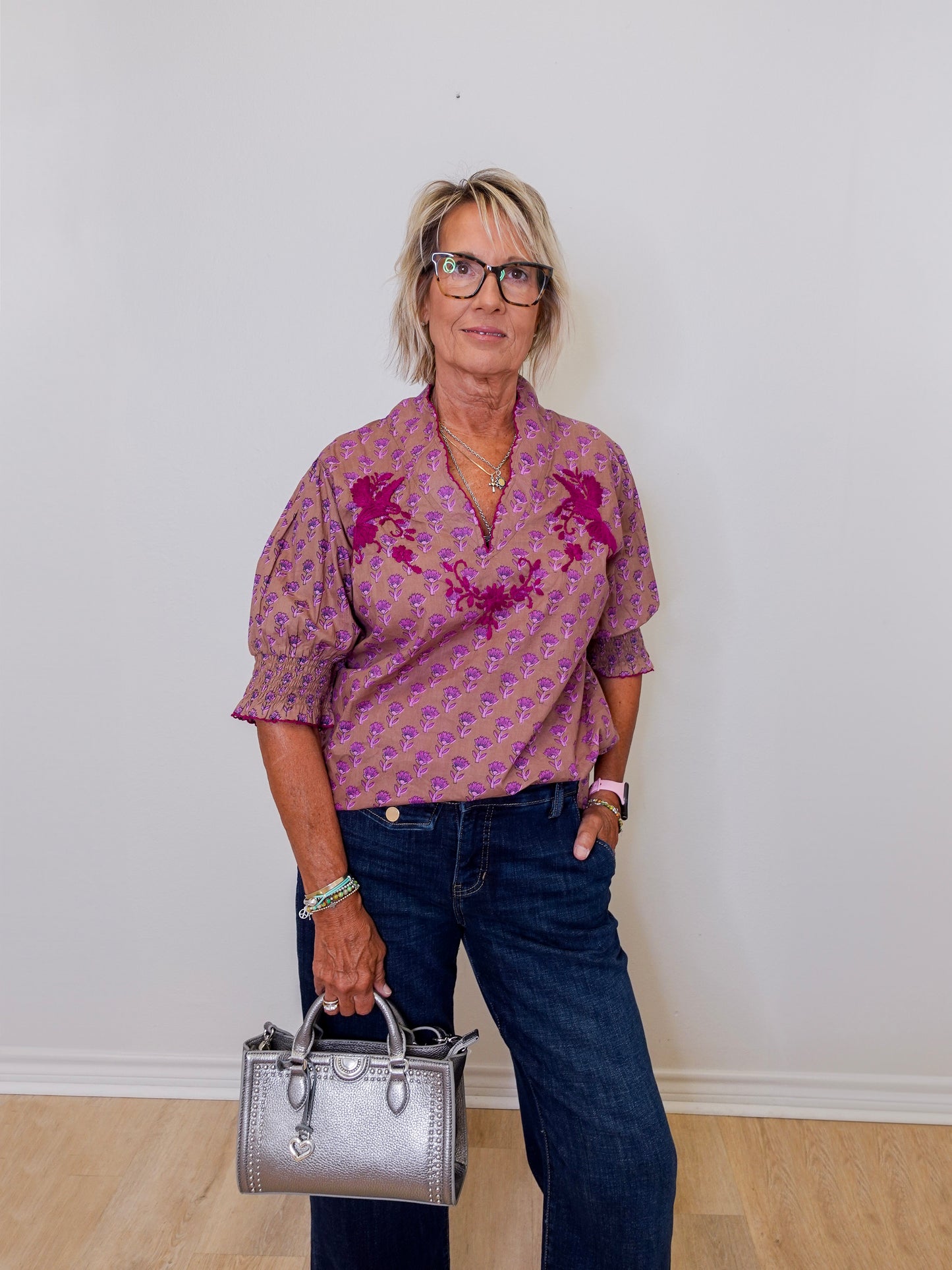 Woman wearing a patterned shirt and jeans, holding a silver handbag against a white wall.