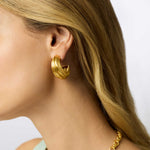 Gold hoop earring worn by a person with blonde hair on a light background