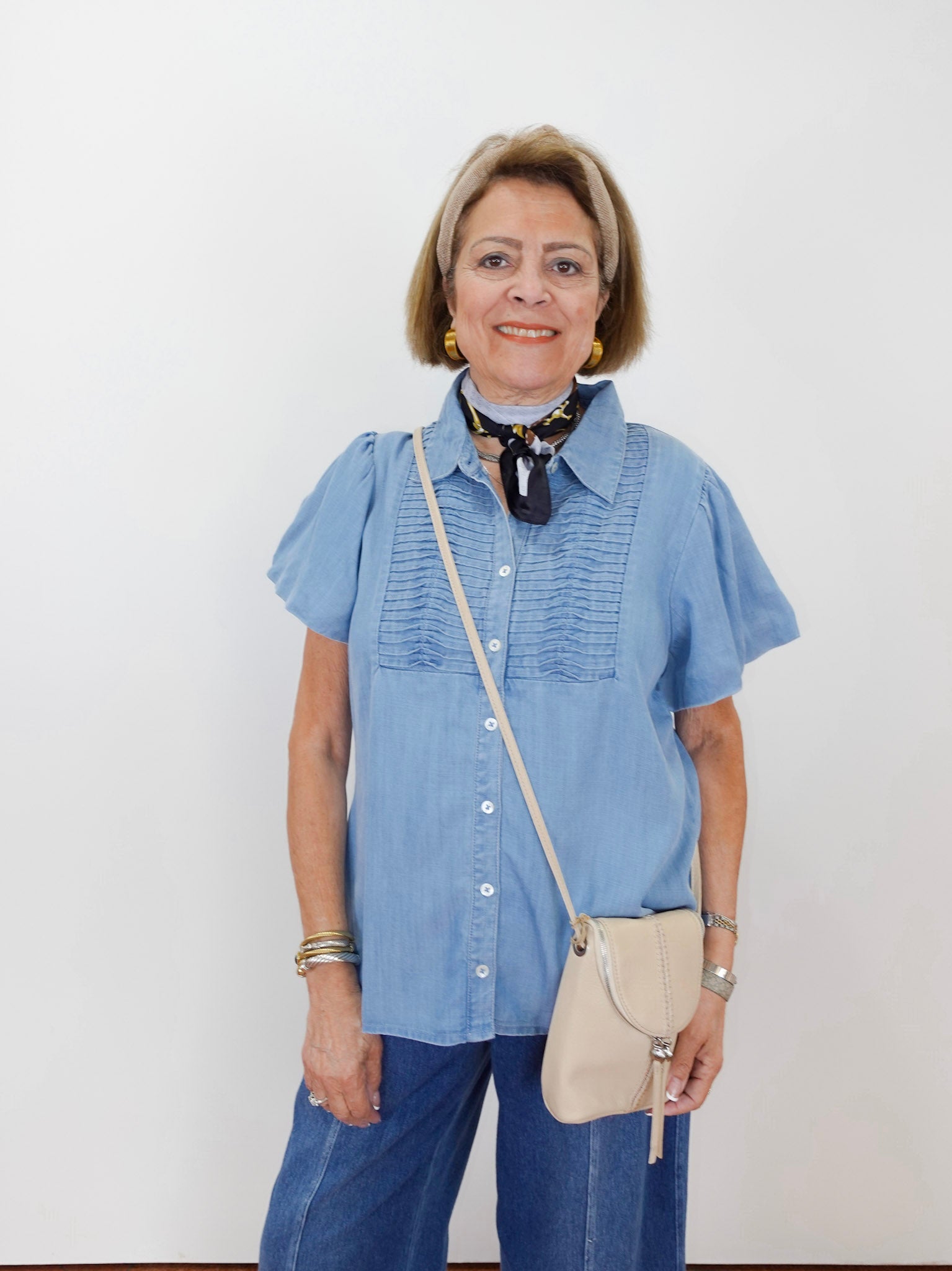 Woman wearing a blue shirt and jeans with a beige crossbody bag against a white background