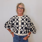 Person wearing a black and white patterned cardigan with blue jeans against a plain background