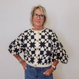 Person wearing a black and white patterned cardigan with blue jeans against a plain background