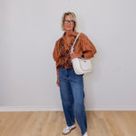 Woman wearing a patterned shirt, jeans, and white sneakers with a white handbag against a plain background.