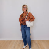 Woman wearing a patterned shirt, jeans, and white sneakers with a white handbag against a plain background.