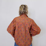 Person wearing a patterned brown shirt and blue jeans against a white wall