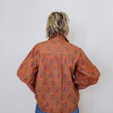 Person wearing a patterned brown shirt and blue jeans against a white wall