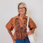 Woman wearing a patterned blouse and jeans, holding a white bag against a plain background