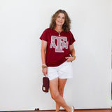 Woman wearing a red t-shirt with a logo and white shorts, standing against a white wall.