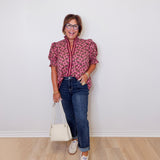 Woman wearing a patterned blouse, jeans, and holding a white bag against a plain background