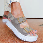 Gray sandal with a thick white sole worn by a person on a patterned floor.