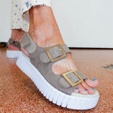 Gray sandal with a thick white sole worn by a person on a patterned floor.