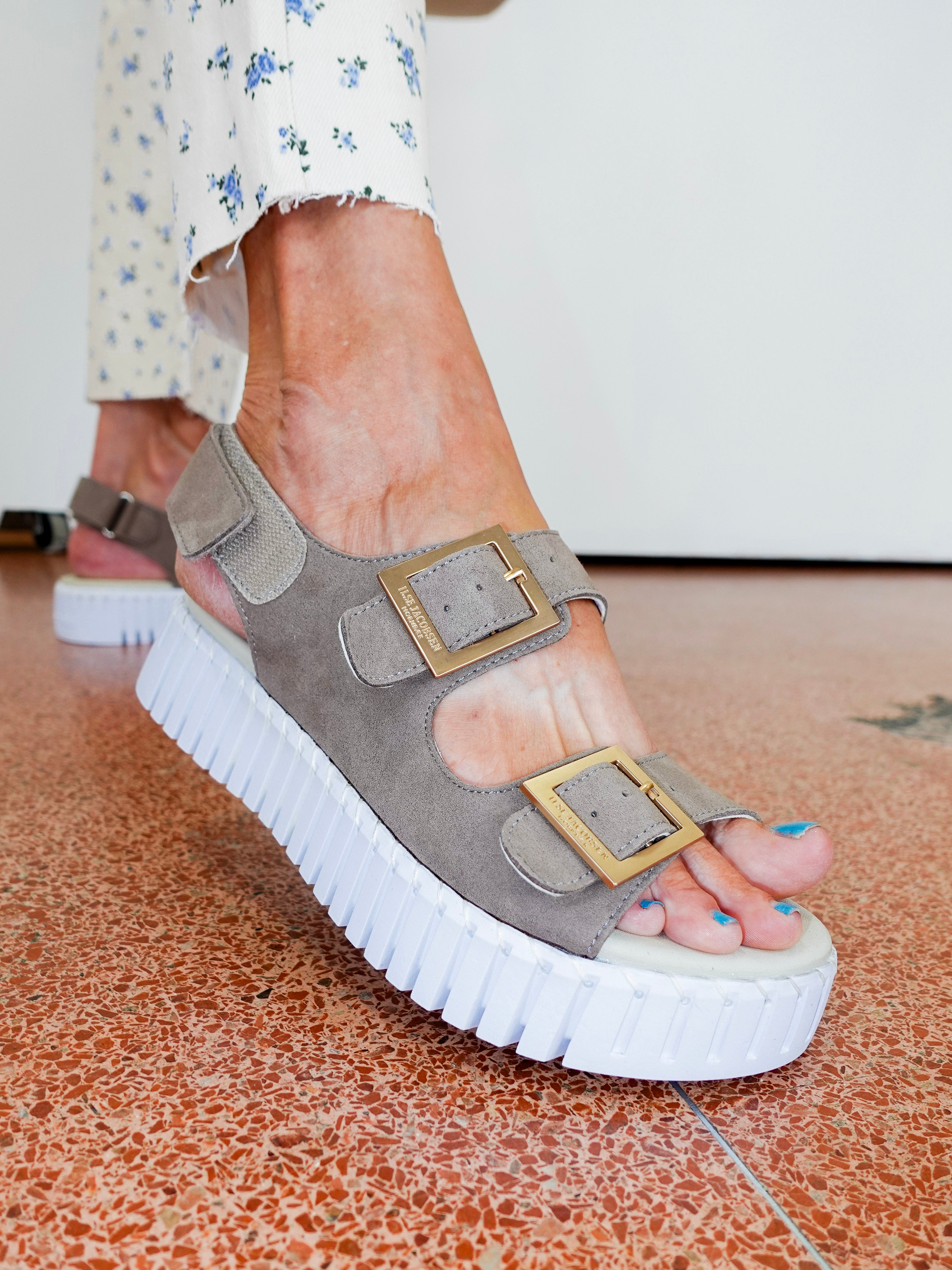 Gray sandal with a thick white sole worn by a person on a patterned floor.