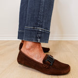 Brown suede loafers worn with rolled-up jeans on a light wooden floor.