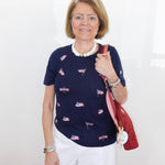 Woman wearing a navy blue top with small flag patterns, holding a red bag against a white background