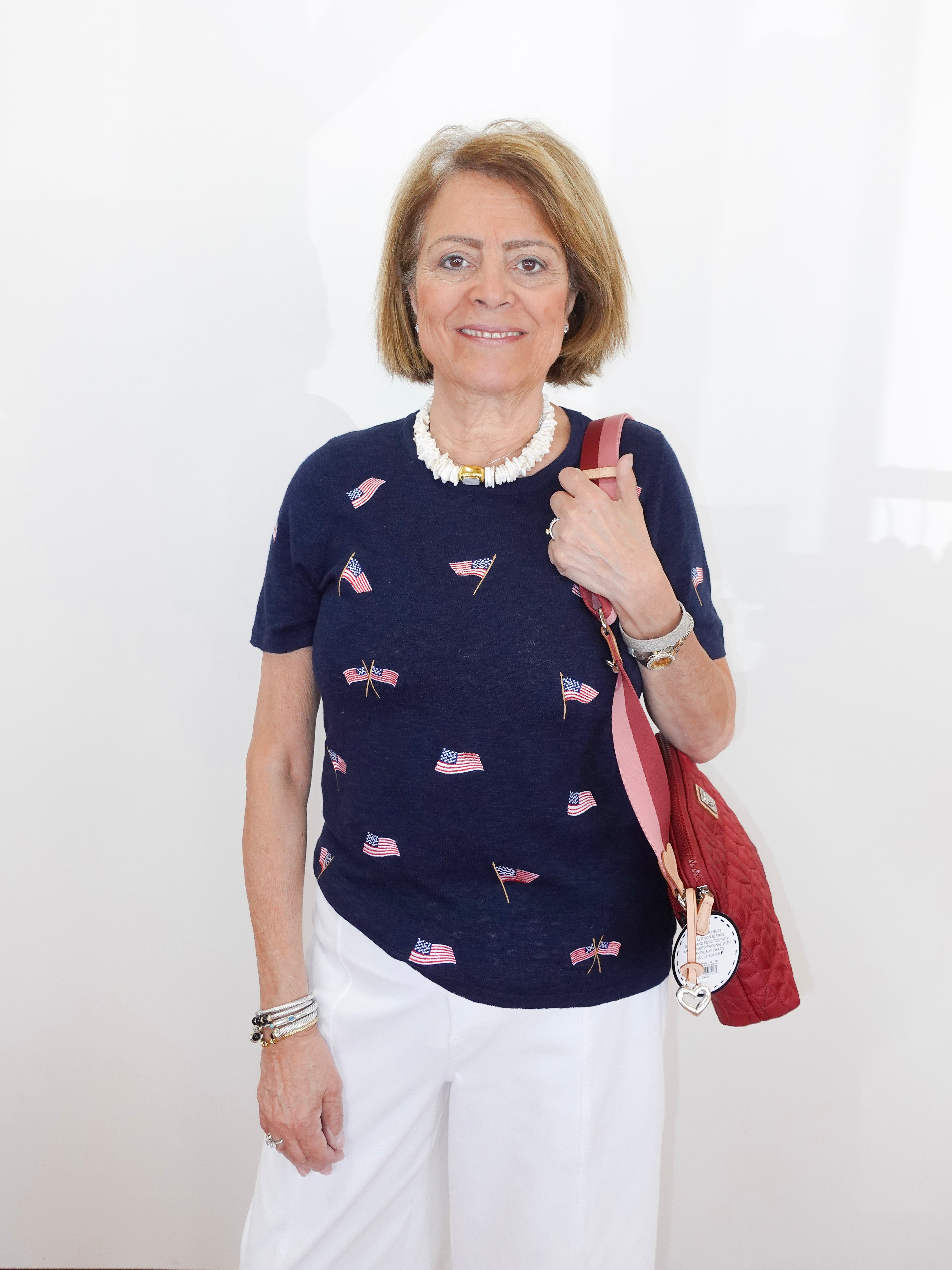 Woman wearing a navy blue top with small flag patterns, holding a red bag against a white background