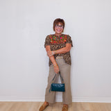 Woman standing against a white wall wearing a patterned top and beige pants, holding a blue handbag.