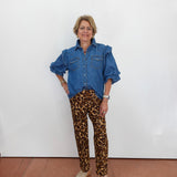 A woman stands against a white wall wearing a blue denim shirt with ruffled shoulders and brown leopard-print pants, paired with beige woven sandals.