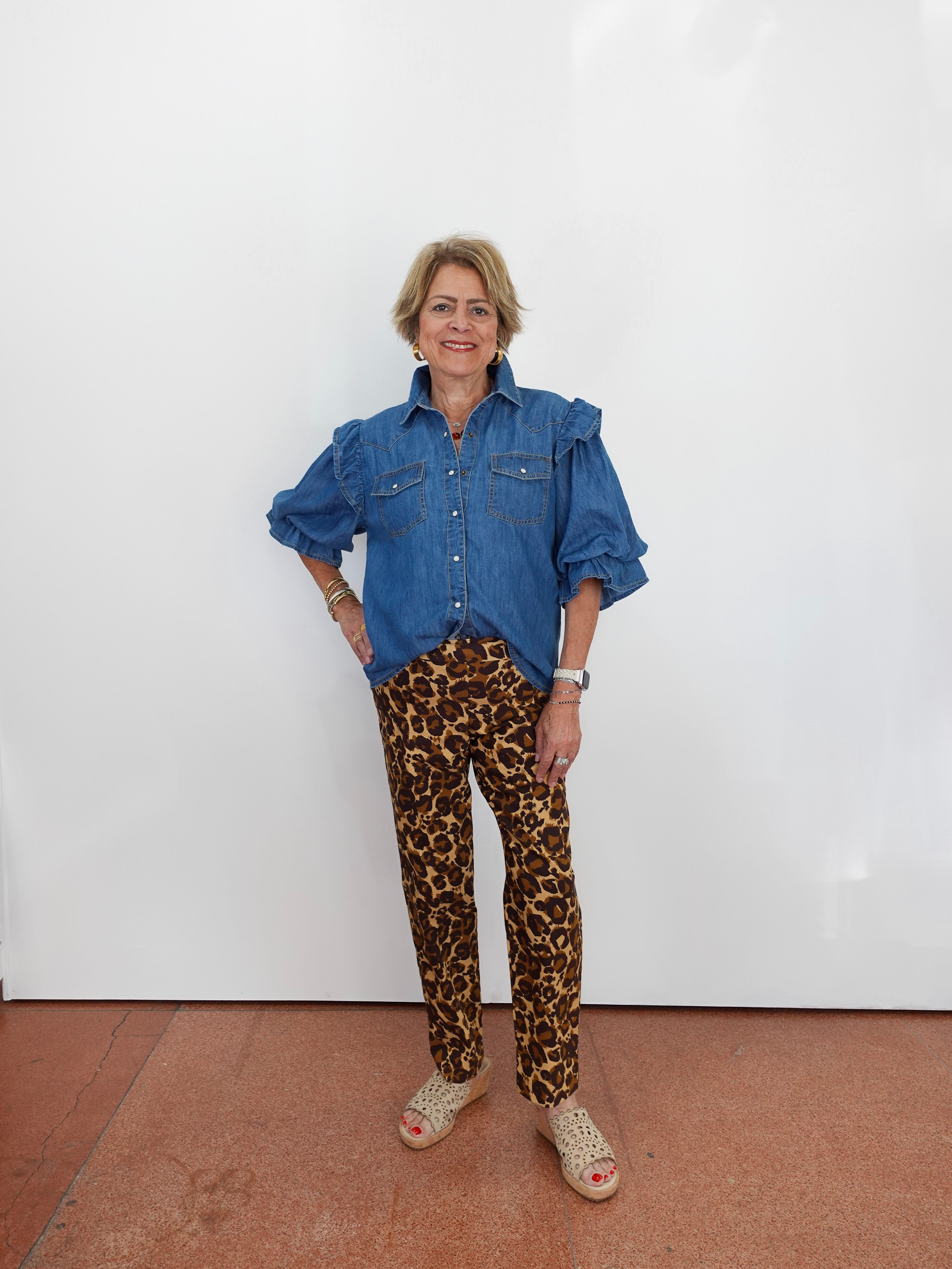 A woman stands against a white wall wearing a blue denim shirt with ruffled shoulders and brown leopard-print pants, paired with beige woven sandals.