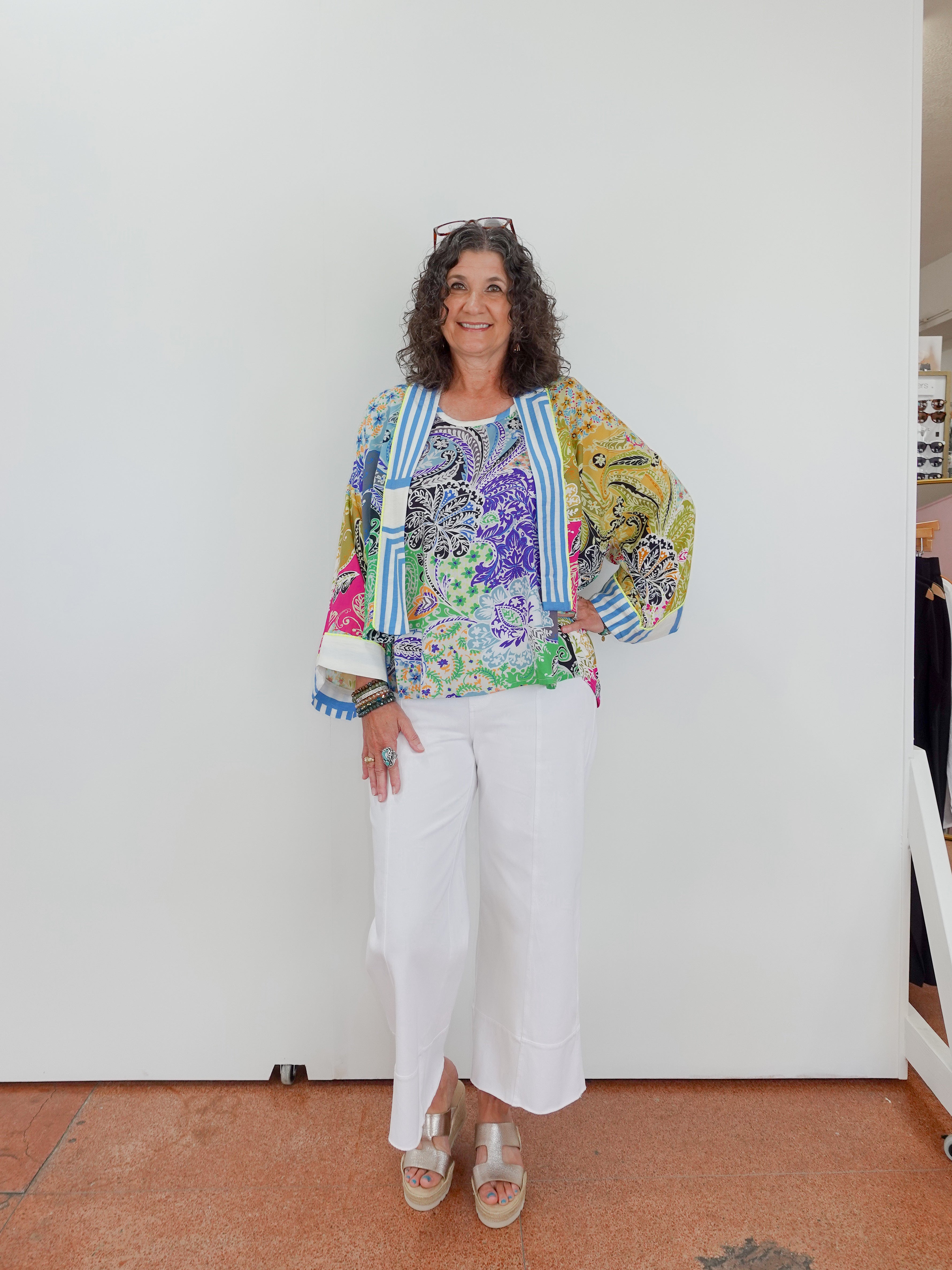 Woman wearing a colorful floral jacket and white pants standing against a white wall.