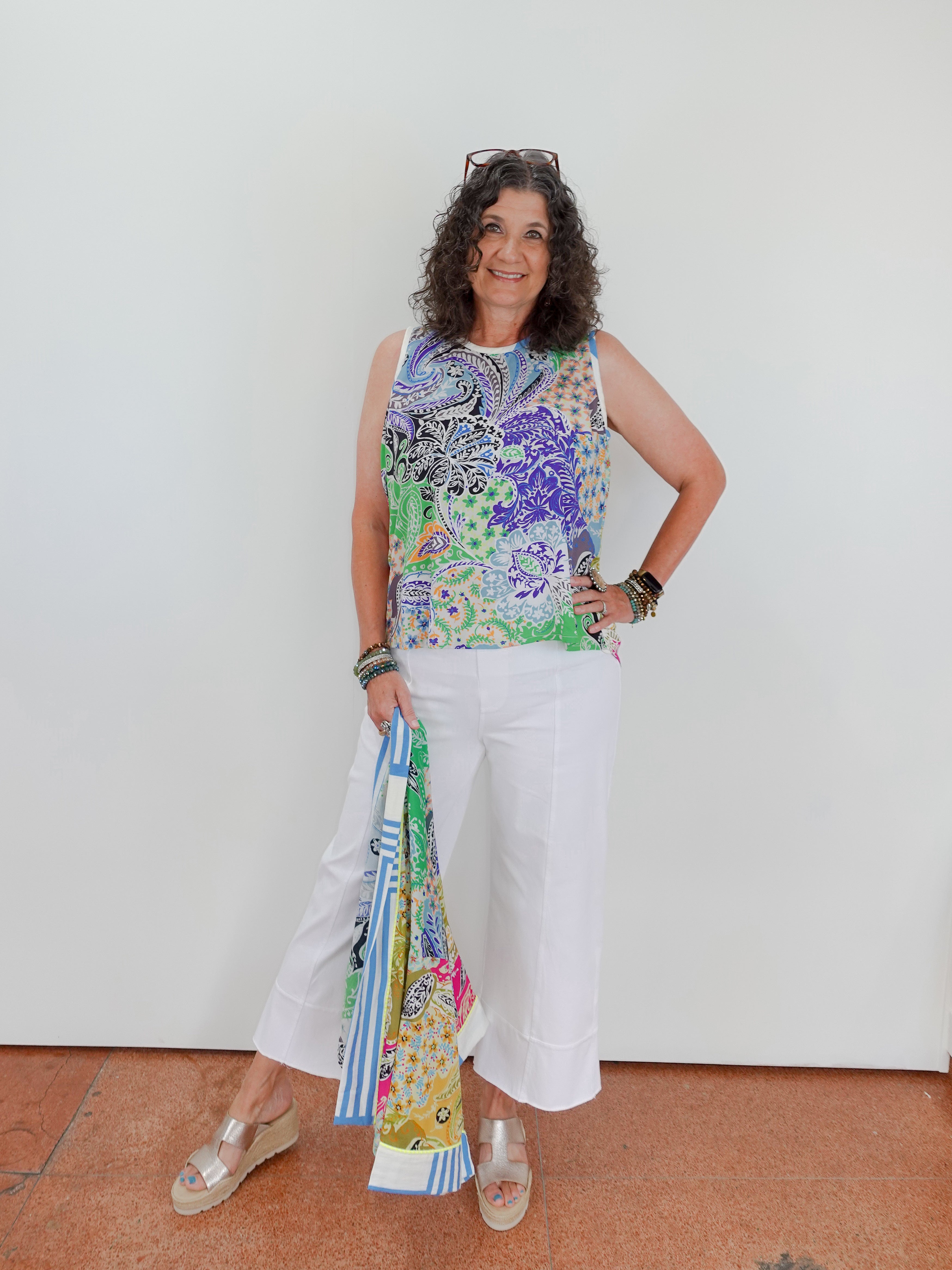 Woman wearing a colorful sleeveless top and white pants, holding a patterned scarf.